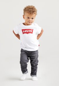 Bela majica s rdečim logotipom 'Levi's', kombinirana z sivimi jeans hlačami. Otrok stoji z rokami za hrbtom, nosi bele nogavice.