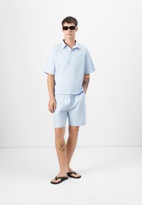 Lyseblå kortermet skjorte med lomme og matchende shorts. Stoffet har et teksturert mønster. svarte solbriller og flip-flops fullfører stilen.