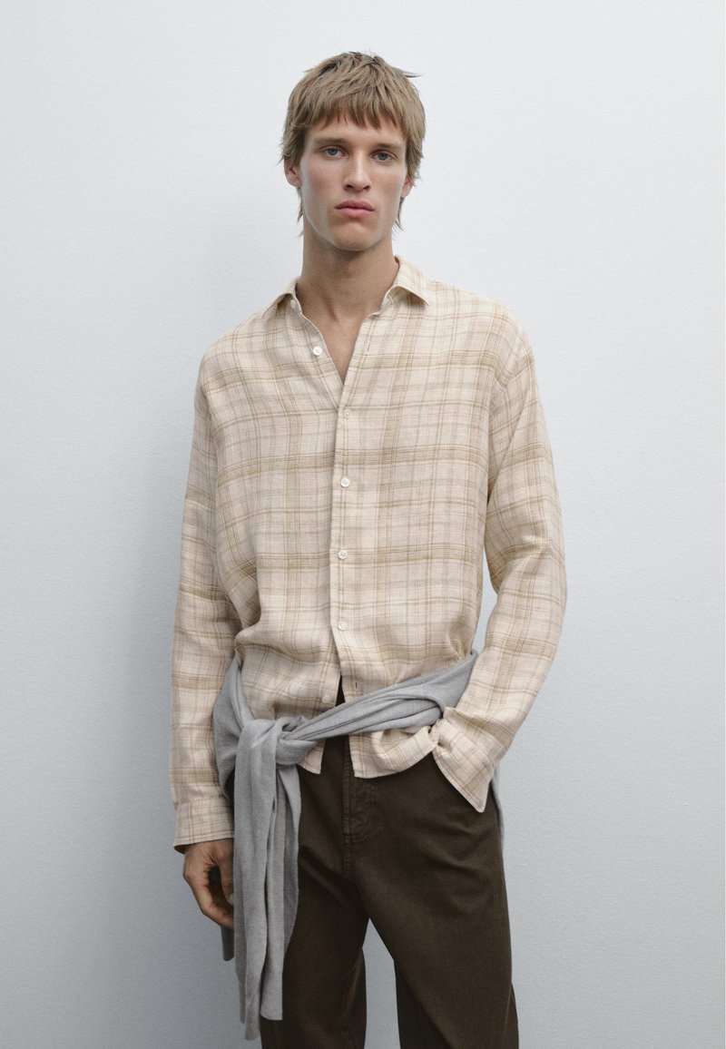 Camicia a quadri beige in tessuto leggero, maniche lunghe, con bottoni, abbinata a pantaloni marroni scuri; tessuto grigio legato in vita.