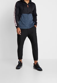 Sort og marineblå lynlås hættetrøje med hætte, der har kontrasterende paneler og logo-detaljer. Sammen med sorte tapered joggers og sneakers.