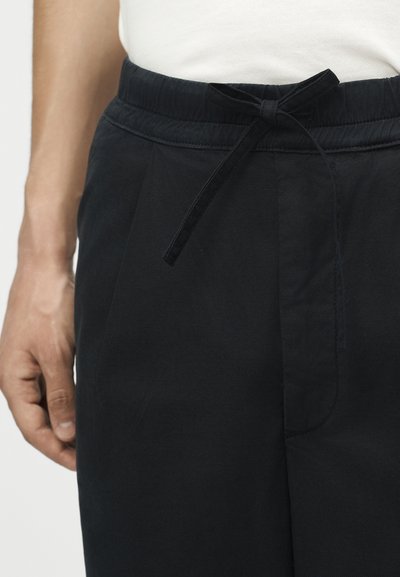 Άνδρας που φοράει μαύρο παντελόνι με ελαστική μέση και δεμένη κορδόνι, συνδυασμένο με λευκό πουκάμισο μέσα στην παντελόνα.