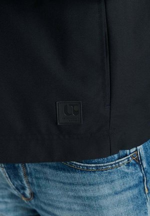 Giacca in tessuto nero con finitura liscia. Presenta una piccola patch rettangolare nera con il logo sull'orlo, abbinata a jeans di denim blu.