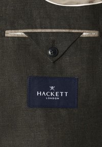 Dunkelgrauer, strukturierter Jackeninnenraum mit beigem Besatz, schwarzer Knopfverschlusss und einem marineblauen Etikett mit der Aufschrift "Hackett London" und gekreuztem Fledermaus-Logo.