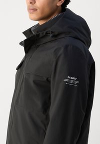 Chaqueta impermeable negra con capucha, que cuenta con un bolsillo frontal y un logo en la manga. Forro interior texturizado visible alrededor del cuello.