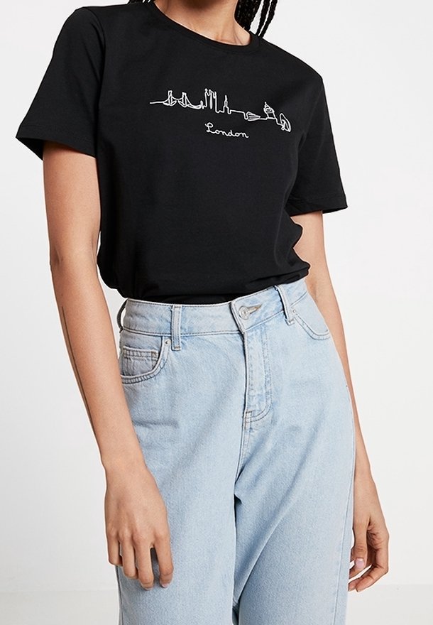 Personne portant un t-shirt noir avec un motif blanc représentant la silhouette de Londres et un jean taille haute bleu clair sur un fond uni.