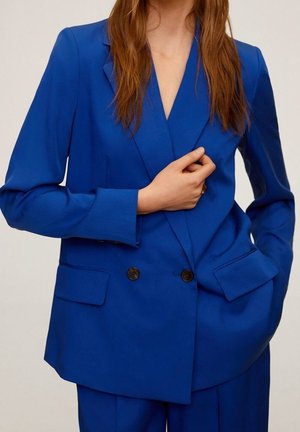 Femme portant un blazer bleu roi à double boutonnage et un pantalon assorti, une main tenant le blazer près de la poitrine et l'autre dans une poche.