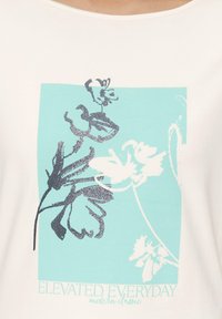 Vitt t-shirt med en turkos grafik som har blommönster i silver och vitt, samt texten "ELEVATED EVERYDAY modern classic" längst ner.