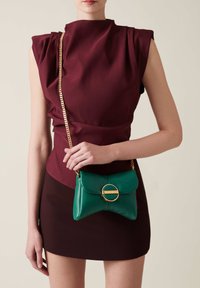 Green leather crossbody bag, rounded shape, gold circular hardware, and chain strap. Worn with a fitted burgundy top and skirt.