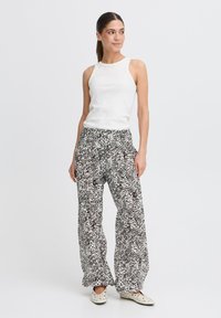 Haut sans manches blanc, pantalon ample à motifs noir et blanc, chaussures décontractées avec un design moucheté, se tenant sur un fond clair.