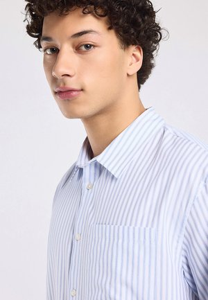 Jonge man met krullend haar die een lichtblauw-wit gestreept overhemd met knopen draagt en iets links naast de camera kijkt.