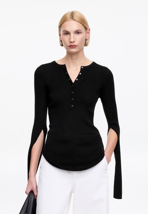 Kobieta o krótkich blond włosach nosi czarną prążkowaną bluzkę z długim rękawem z przodu zapinaną na guziki i z rozciętymi rękawami, połączoną z białymi spodniami, trzyma czarną torbę.