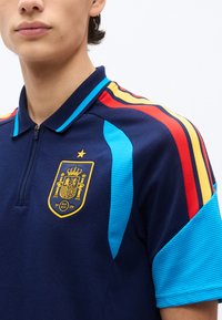 Artimos planas žmogaus, dėvinčio tamsiai mėlyną sportinį marškinėlį su raudonomis, geltonomis ir mėlynomis juostelėmis ant peties ir geltonu emblemų su karūna ant krūtinės.