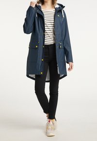 Mörkblå regnjacka med huva, framsidan med dragkedja, gula knappar och två bröstfickor. Matchad med en randig skjorta och svarta byxor.