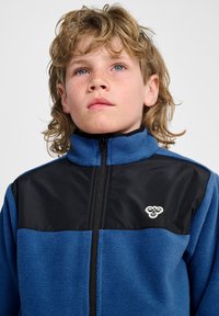 Blauwe fleecejas met zwarte stof op de schouders, hoge kraag en ritssluiting aan de voorkant. Heeft een klein wit logo op het borstgedeelte.