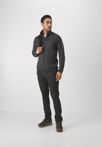 Veste zippée grise avec col montant, en tissu texturé et poches latérales, associée à un pantalon noir et des chaussures robustes, portée par un homme.