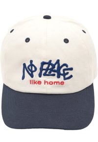 Keps med en krämfärgad front och en marinblå skärm, broderad text "NO FLAG like home" i blått och rött, strukturerad form och tygmaterial.