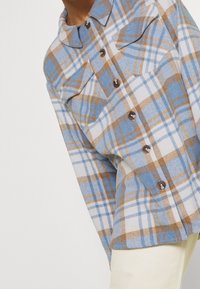 Camicia a quadri blu, bianca e marrone con tasche sul petto indossata su pantaloni chiari.