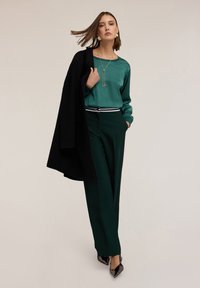 Top verde in satin, pantaloni verdi a vita alta con una cintura a righe bianche e nere, cappotto nero gettato sulla spalla e scarpe nere a punta.