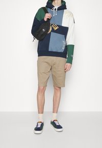 Sweat à capuche avec patches multicolores (marine, vert, bleu clair, blanc) ; shorts beige ; sac bandoulière noir ; baskets bleues et vertes.