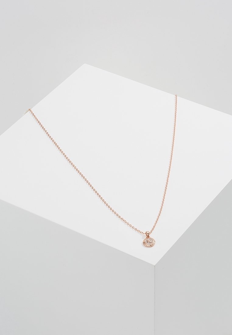 Collier délicat en chaîne d'or rose avec un petit pendentif circulaire doté d'une pierre précieuse claire, exposé sur un piédestal géométrique blanc.