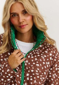 Brązowa kurtka w cętki z zielonym wnętrzem i zamkiem błyskawicznym, o miękkiej fakturze. Widoczny jest naszyjnik z kolorowych koralików.