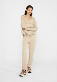 Sweat à capuche beige avec un col en V et un cordon, assorti à un pantalon beige avec des accents sur les côtés. Chaussures de sport marron et crème.