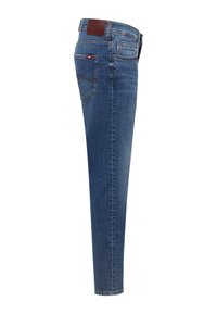 Jean en denim bleu coupe ajustée avec poche arrière, écusson en cuir de la marque sur la ceinture, et détails de couture visibles sur un fond blanc.