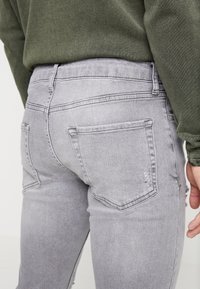 Jeans de mezclilla gris claro vistos desde atrás, con un ligero desgaste, dos bolsillos traseros y una textura ligeramente desgastada. Acentuados por una blusa verde.