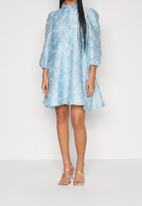 Kvinna klädd i en ljusblå blommig brokadklänning med puffärmar och guldhögklackade sandaler, stående mot en neutral bakgrund.