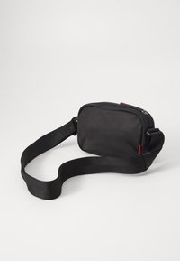 Geantă rectangulară neagră, fabricată din material neted. Prezintă un sistem de închidere cu fermoar, curea ajustabilă și un mic accent roșu. Design compact și minimalist.
