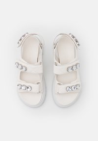 Sandales blanches avec deux sangles réglables ornées de strass clairs décoratifs. La tige douce a une finition texturée, et une semelle blanche épaisse.