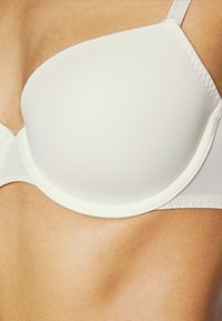 Bra imbottito bianco con tessuto liscio, dotato di spalline regolabili e dettagli di cucitura. Il design include un ferretto curvo per offrire supporto.