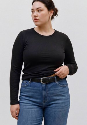 Femme aux cheveux foncés attachés en chignon portant des boucles d'oreilles créoles argentées, un haut noir à manches longues, un jean bleu et une ceinture noire, regardant sur le côté.