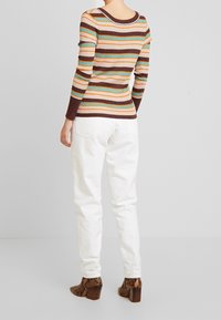 Person som bär en långärmad randig tröja, vita byxor och bruna ormskinnsankelboots, stående vänd bortåt mot en neutral bakgrund.