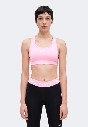 Mladá osoba s krátkymi tmavými vlasmi nosí ružovú športovú podprsenku Nike a čierne legíny s ružovým pásom Nike Pro a stojí na jednofarebnom pozadí.