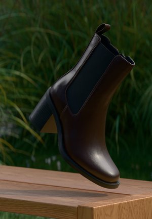 Stivaletto alla caviglia in pelle marrone scuro con pannello laterale elastico e tacco a blocco sospeso sopra una panchina di legno con sfondo di erba.
