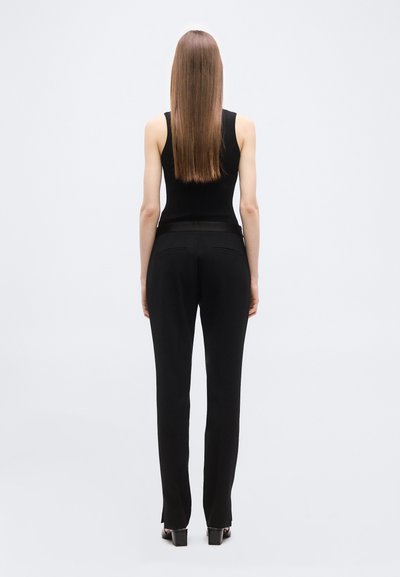 Femme aux longs cheveux raides, vue de dos, portant un débardeur noir, un pantalon noir et des chaussures à talons noires sur un fond uni.