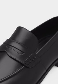 Mocassin en cuir noir avec couture moc toe et détail de sangle sur le dessus, exhibant une texture lisse et un design élégant.