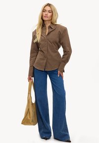 Chemise à manches longues marron à carreaux avec une silhouette ajustée, associée à un jean large bleu taille haute et un sac fourre-tout beige.