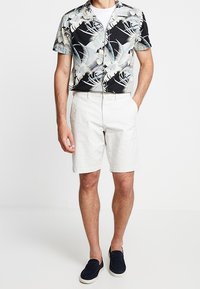Camisa de manga corta negra con estampado floral, hecha de un tejido ligero, combinada con pantalones cortos beige claros. Llevada con zapatos de slip-on azul marino.