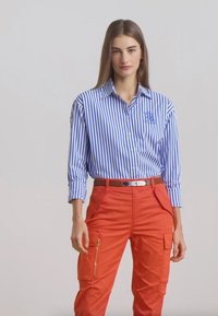 Blauw-wit gestreept shirt met een kraag, gecombineerd met feloranje cargobroek met ritsen en zakken, gedragen met een bruine riem.