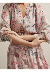 Femme croisant les bras, portant un chemisier transparent à motifs floraux aux tons roses, verts et rouges pâles, avec des manches retroussées et des boutons sur le devant.