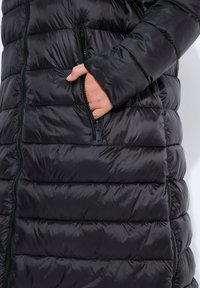 Abrigo de plumas negro con diseño acolchado, textura suave y un bolsillo con cremallera. Presenta un cuello alto y puños ajustables para mayor calidez.