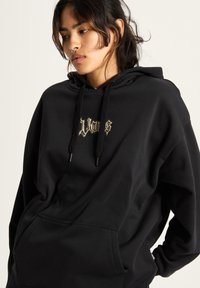 Schwarzer oversized Hoodie aus weichem Stoff mit einer frontalen Kängurutasche und einem grafischen Logo in hellen Farben auf der Brust.