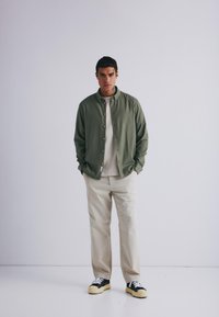 Camicia verde oliva con bottoni, t-shirt beige, pantaloni color crema e sneakers nere con lacci bianchi. Tessuto morbido, vestibilità casual, design semplice.