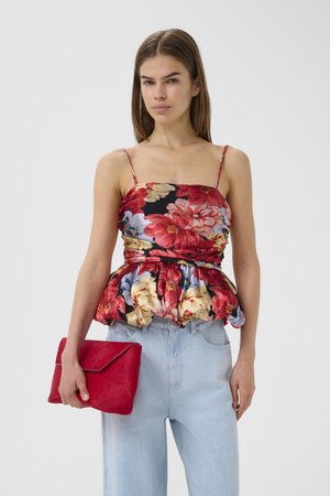 Frau trägt ein geblümtes Peplum-Top mit dünnen Trägern, hellblaue Jeans und hält eine rote Clutch vor einem schlichten Hintergrund.