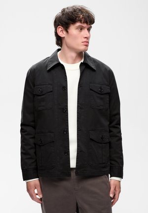Jeune homme portant une veste noire boutonnée sur un pull blanc, regardant vers sa droite sur un fond clair uni.
