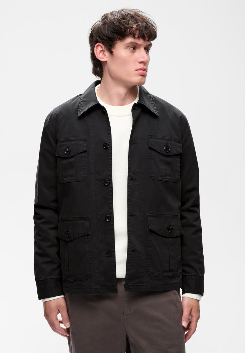 Jeune homme portant une veste noire boutonnée sur un pull blanc, regardant vers sa droite sur un fond clair uni.