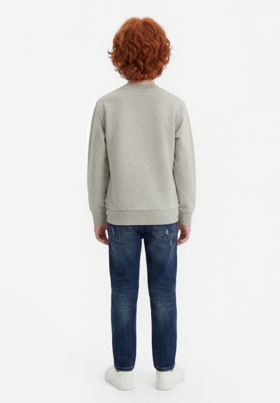 Dziecko z kręconymi rudymi włosami, ubrane w jasnoszarą bluzę, niebieskie dżinsy i białe trampki, stojące tyłem na białym tle.