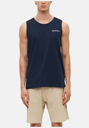 Mann trägt ein marineblaues ärmelloses Shirt mit "Après Surf" Text und beigen, strukturierten Shorts und steht vor einem einfarbigen Hintergrund.